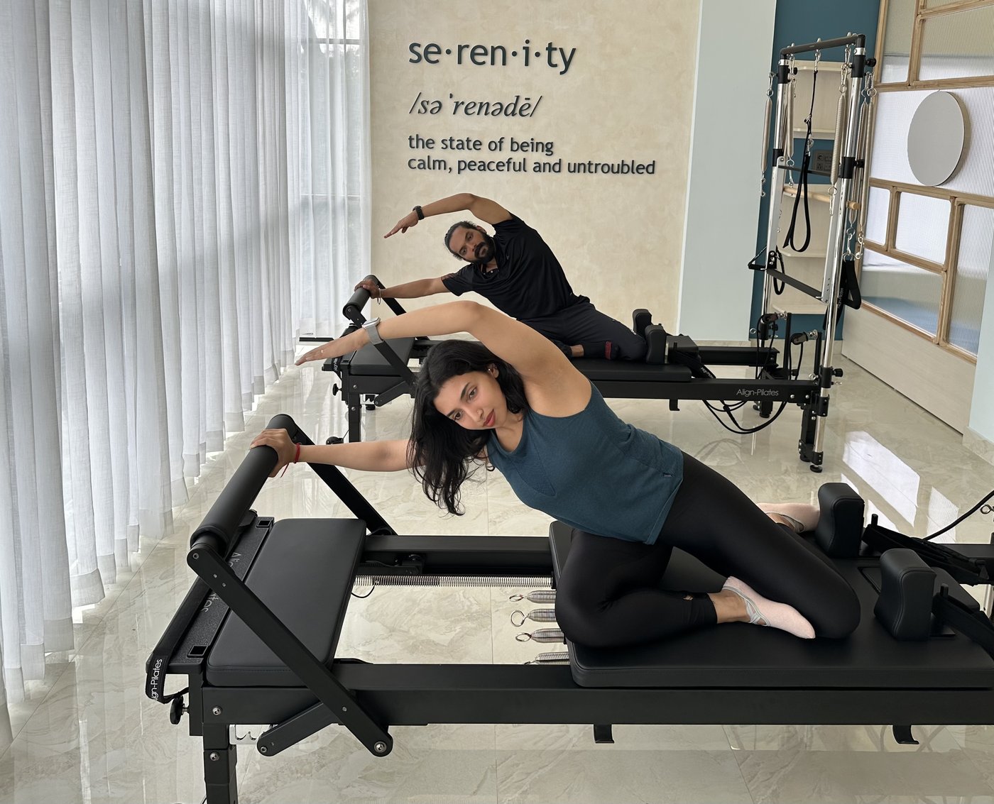 Two instructors doing a side stretch on reformers in front of the Serenity wall